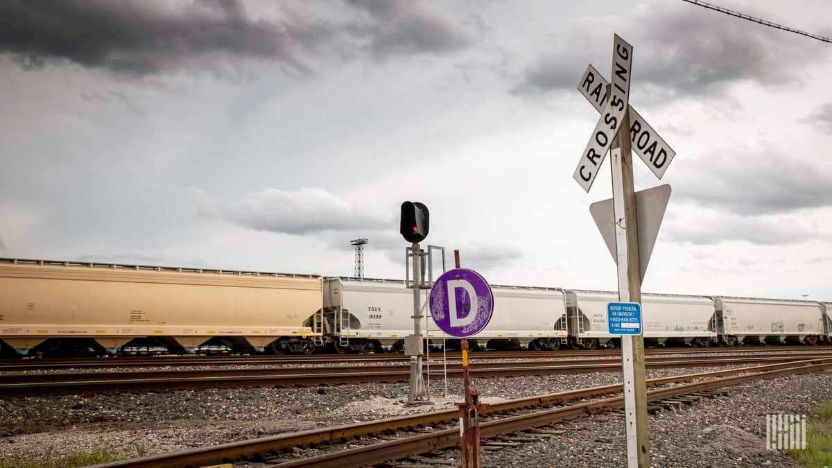 A photograph of a train hauling grain hopper cars. The train is at a railroad crossing.