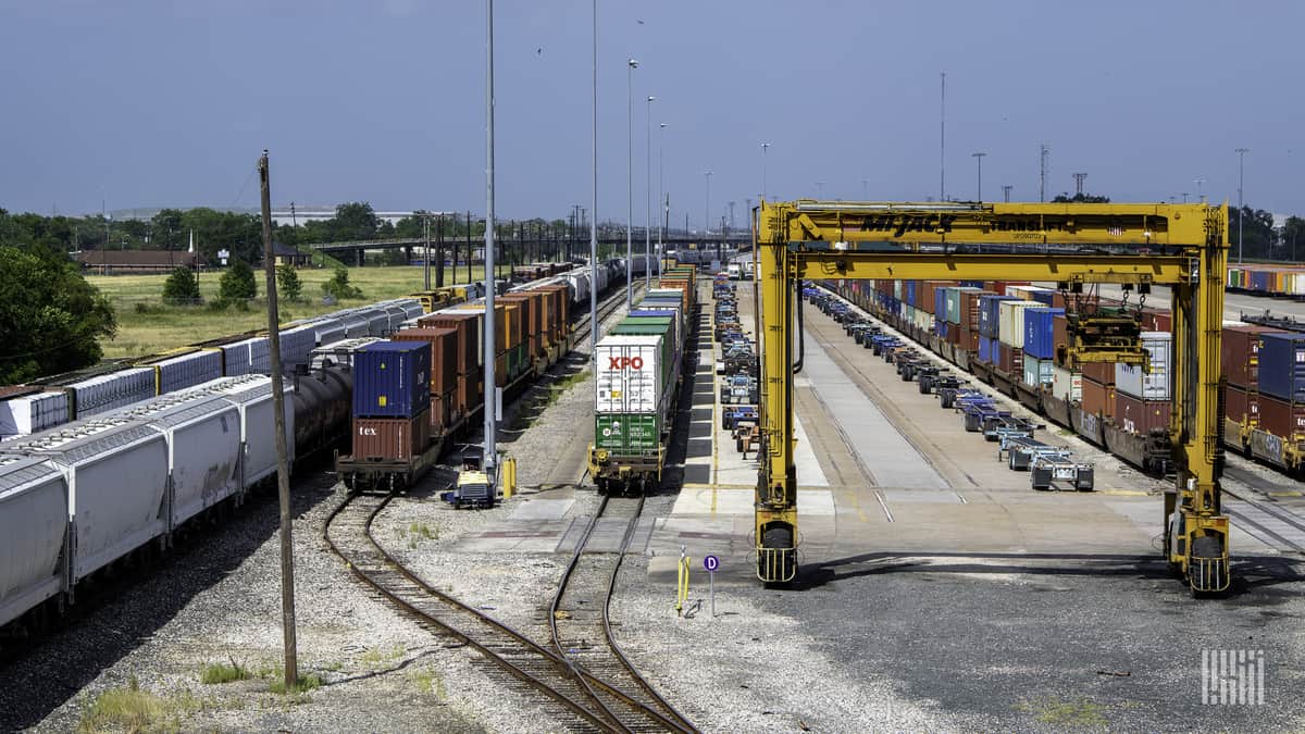 A photograph of a rail yard with several rows of parked intermodal containers and hopper cars.