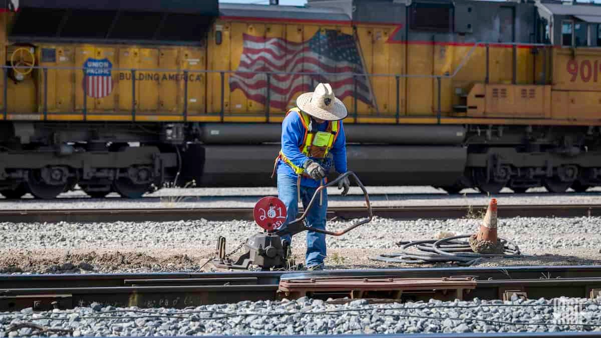 A photograph of a worker fixing track in a rail yard. There is a parked train behind him.