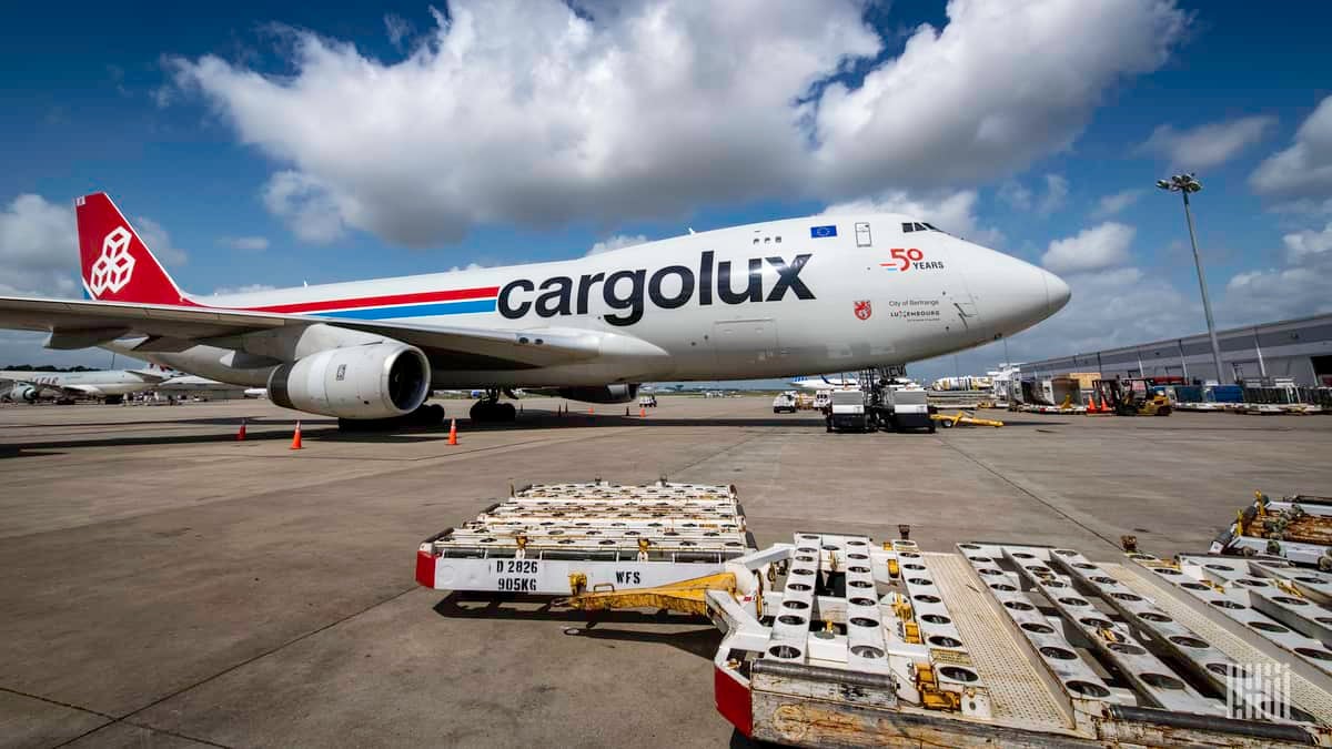 A white cargo jumbo jet with empty pallets on the ground on sunny day. The side of the plane says Cargolux.