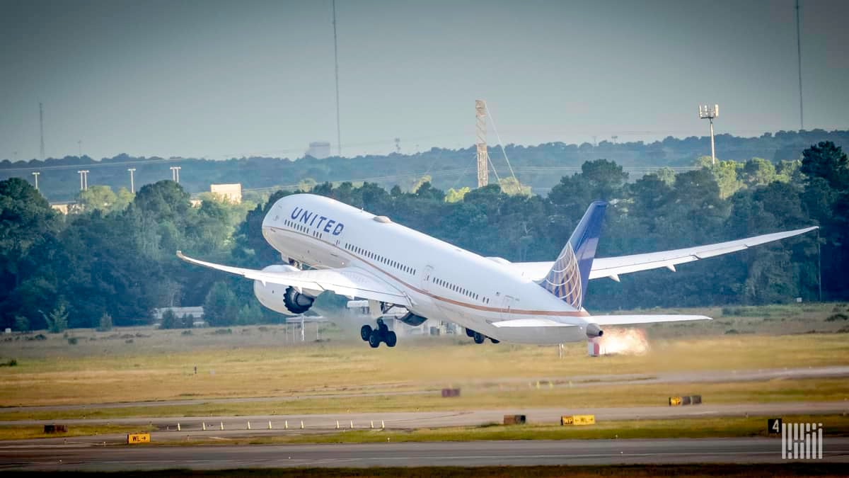 A large white jet with blue tail lifts off from runway on sunny day. View is from behind the plane, to the side a bit.