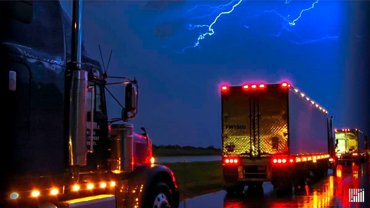 Tractor-trailers heading down highway in the rain, with lightning across the sky.