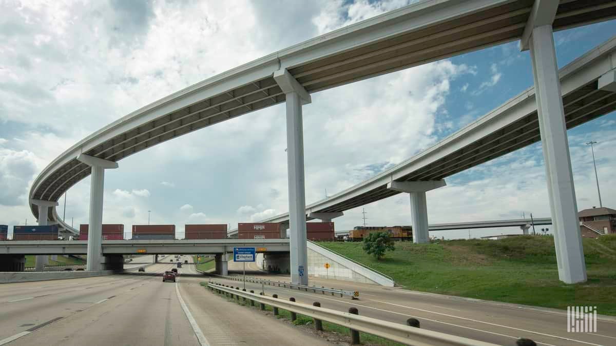 A photograph of a train hauling intermodal containers by a highway overpass.