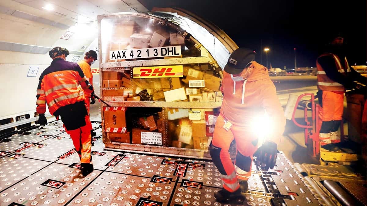 Workers in yellow vests load container in illuminated cargo bay of airplane at night.