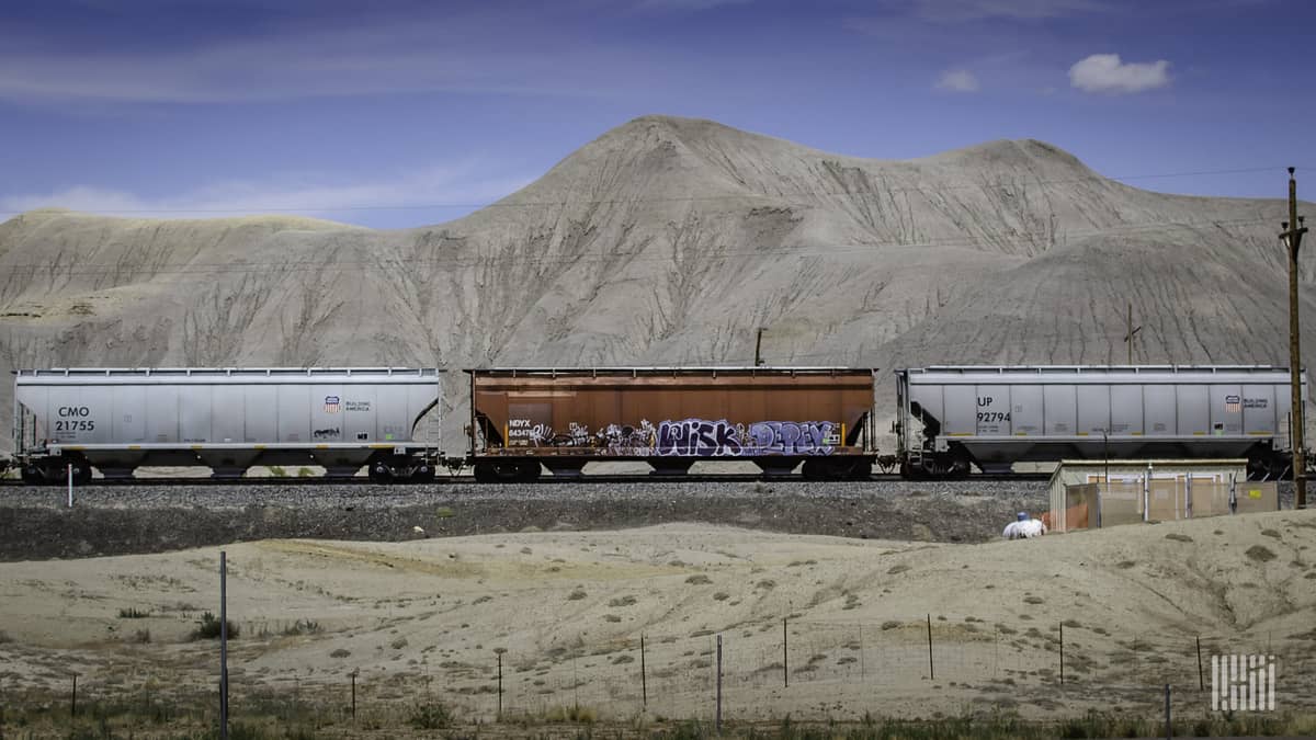 A photograph of grain hopper cars traveling on some railroad track. There is a mountain range in the distance.