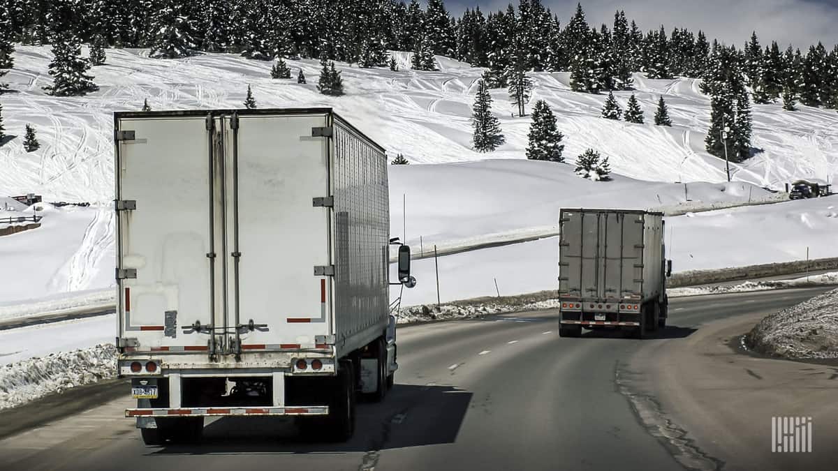 Tractor-trailers on a slick highway with snow in the surrounding mountains.