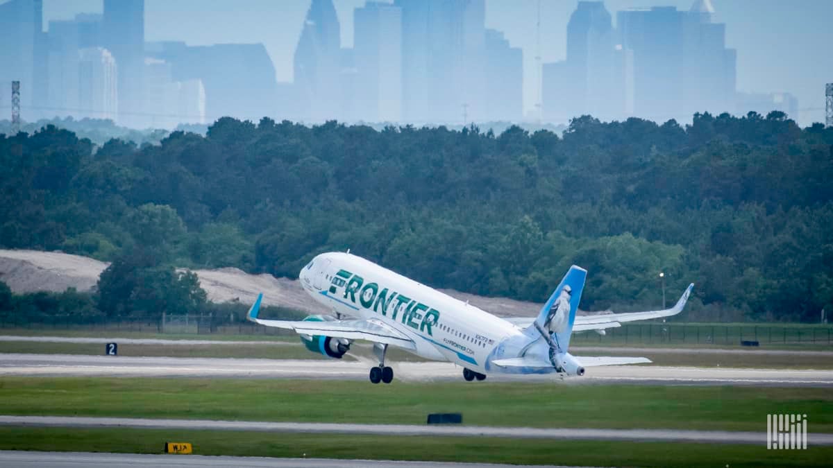 A white jet with light green tail lifts off runway with green trees and city skyline in the background.