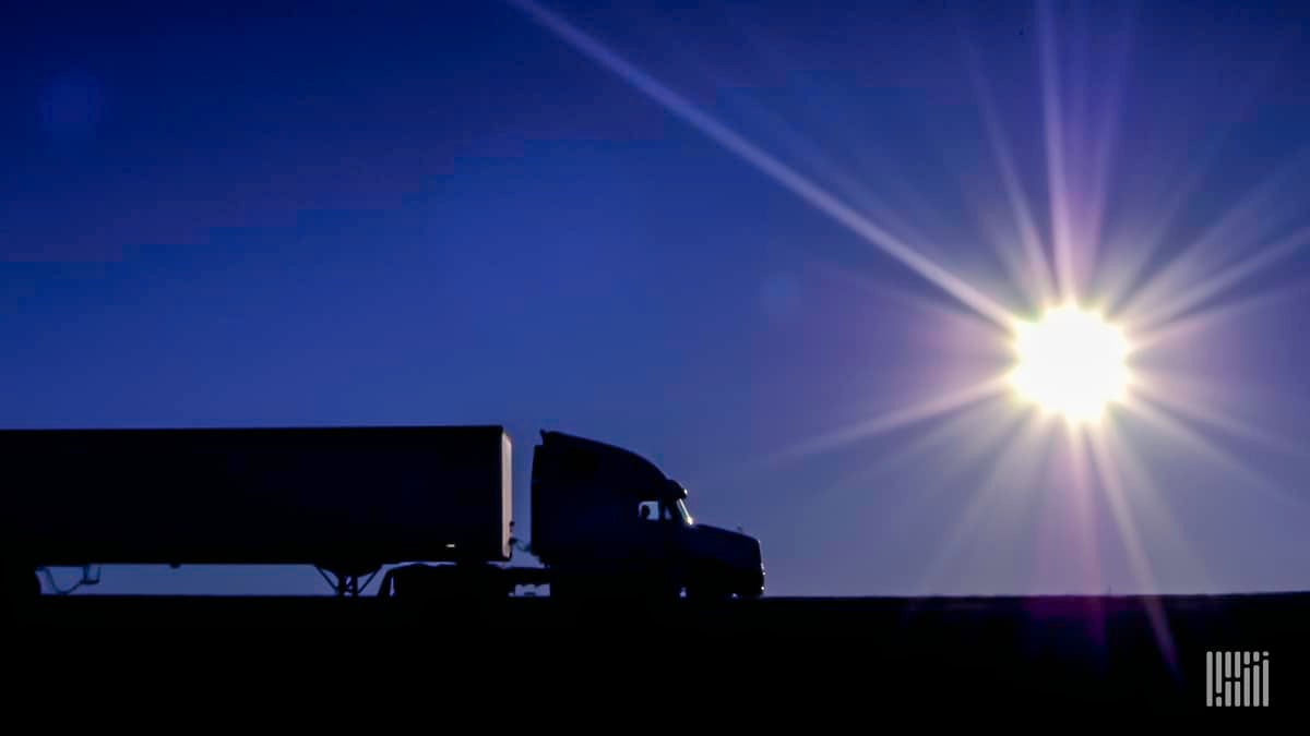 Tractor-trailer heading down a highway at dusk/dawn with bright sun on the horizon.