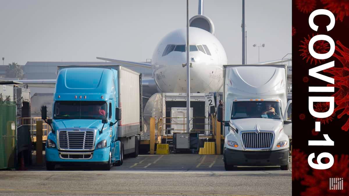 A big white jet flanked by a blue and white truck on each side of nose. COVID-19 banner since both types of transport will be used to deliver coronavirus vaccines.