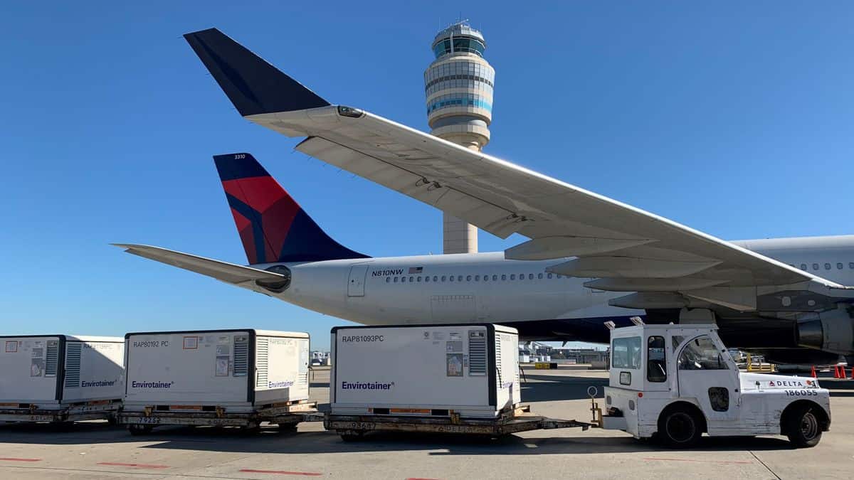 Temperature-controlled airfreight containers on carts being pulled to Delta plane for loading on sunny day. View of aircraft from below wing and towards back half of airplane.