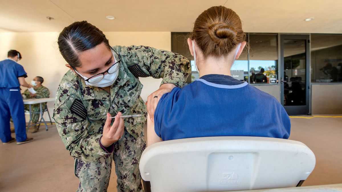 A woman in Army fatigues gives a shot in the arm to another woman with her back facing the camera.