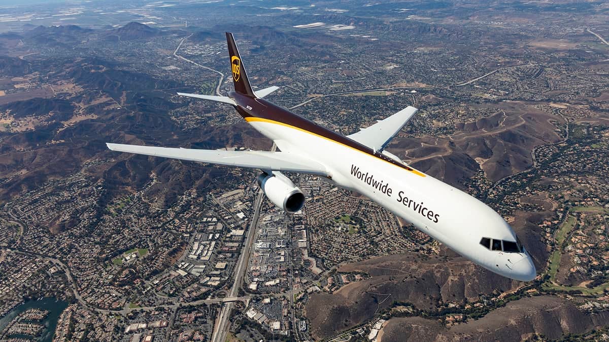 A white UPS cargo jet with brown tail flies over populated area. View is looking down from above aircraft.