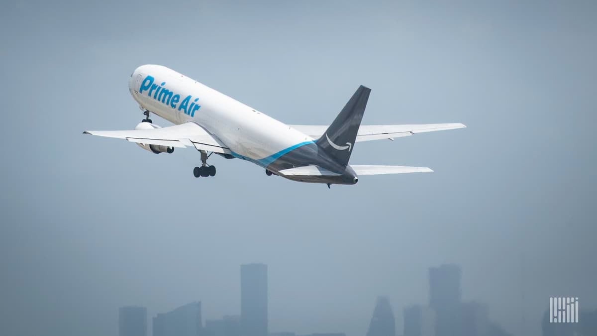 A white Amazon Air jet with blue tail lifts off with city skyline in background.