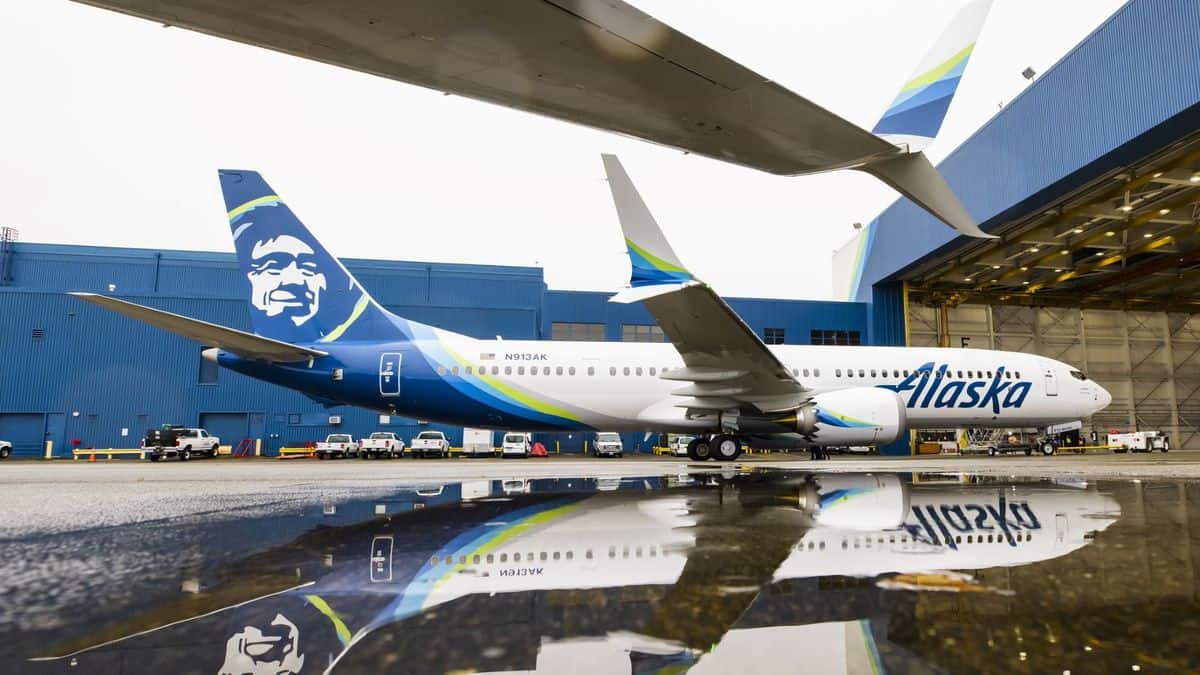 A white 737 MAX with Native Alaskan logo on tail sits in front of a hanger on wet pavement. Ground view from the side.