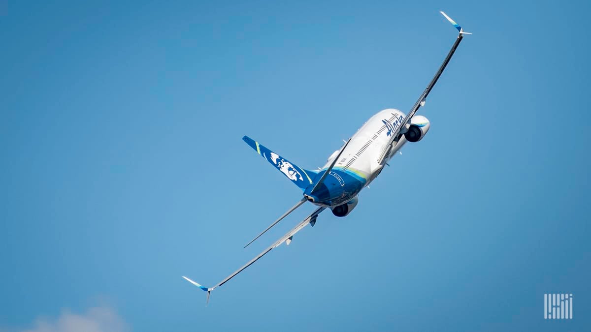 A white Alaska Airlines jet banks to the left during its climb into a blue sky, view from behind.