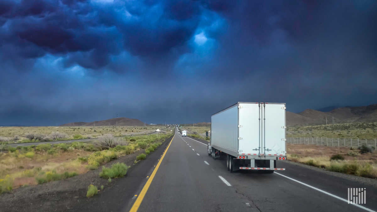 Tractor-trailer heading down highway with thunderstorm cloud across the sky.