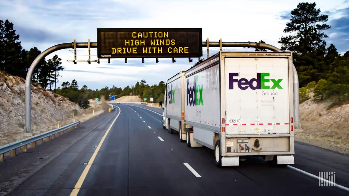 FedEx truck heading down highway with "Caution: High Winds" digital sign ahead.