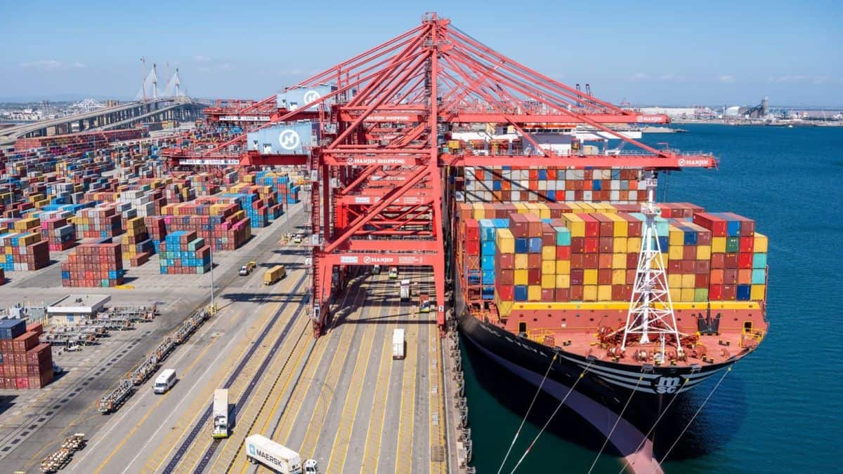 A big container ship stacked with boxes next to the wharf with big cranes and containers stacked on land.