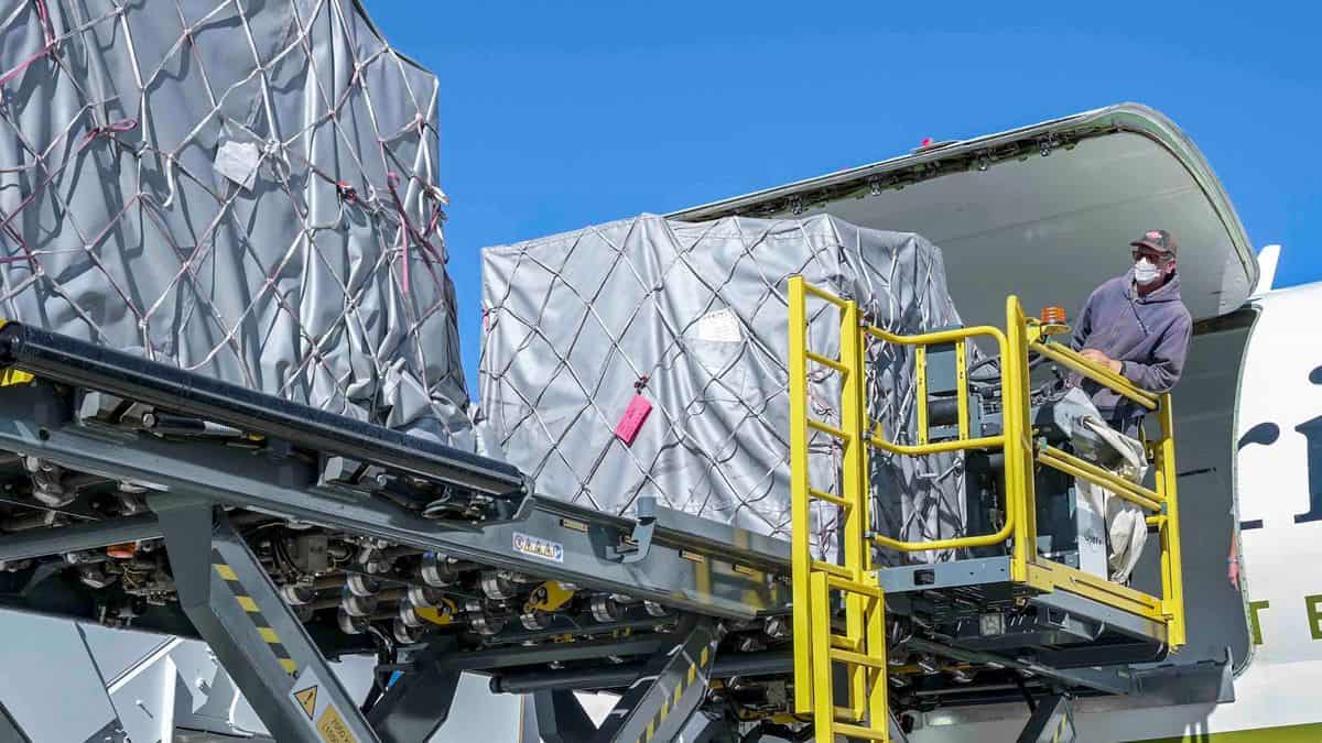 Containers on a lift get loaded into side a big airplane on a sunny day.