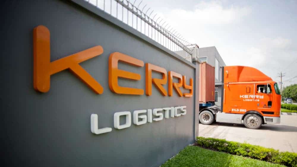 An orange Kerry Logistics truck entering road from Kerry facility next to company sign.