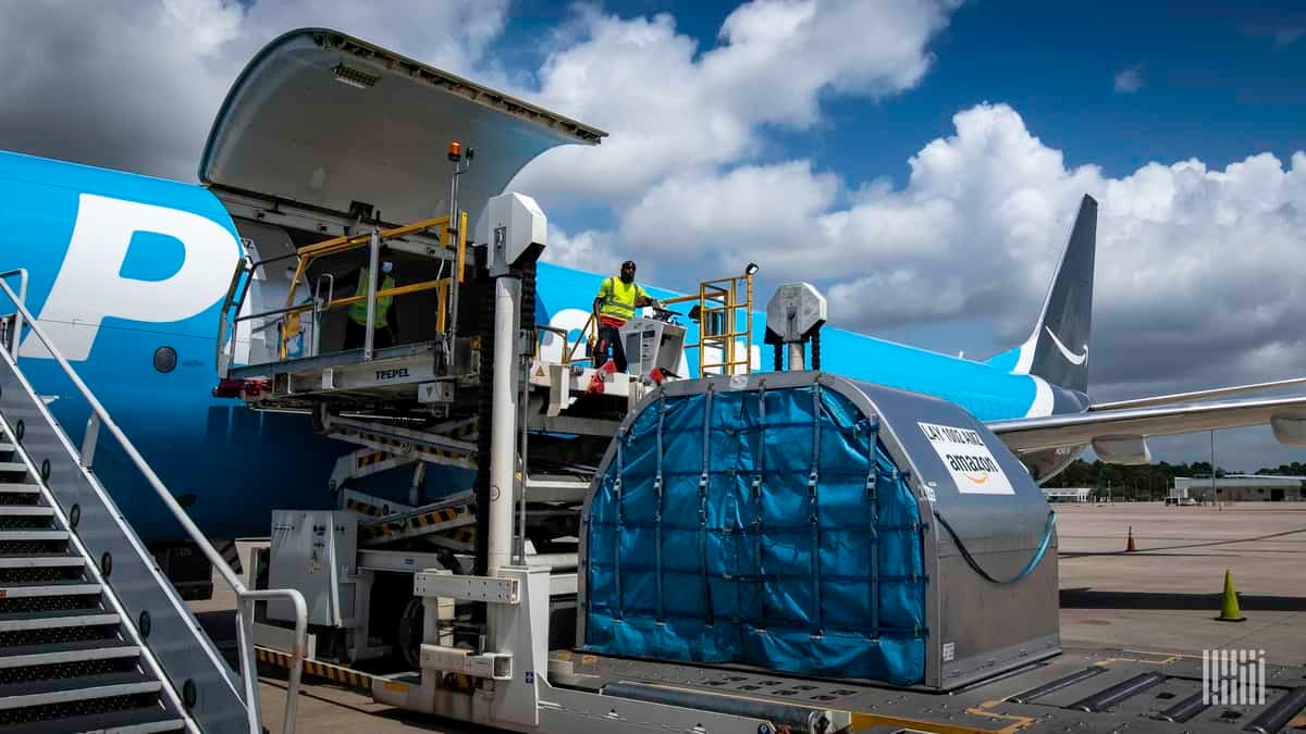 A light blue Amazon plane with side cargo door open and large metal container on the ground ready to be lifted for loading.