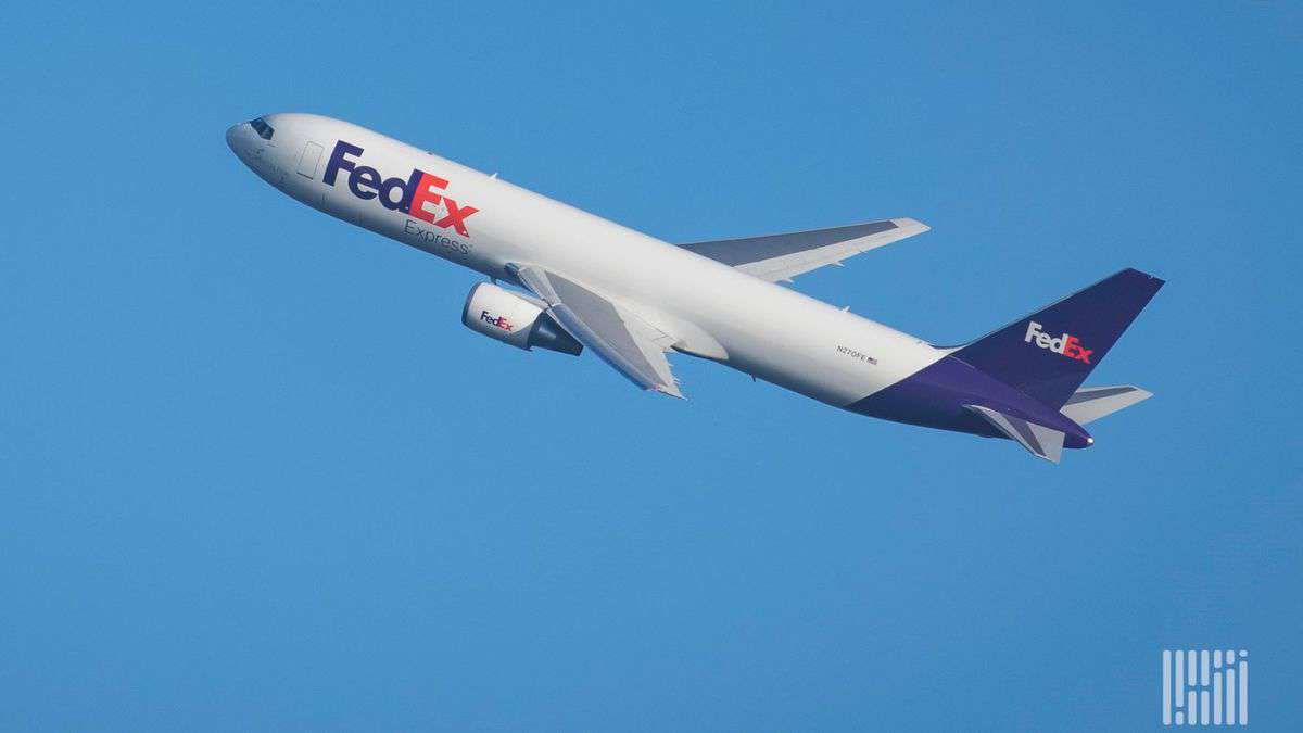 A white FedEx cargo jet with blue tail climbing through a bright blue sky.