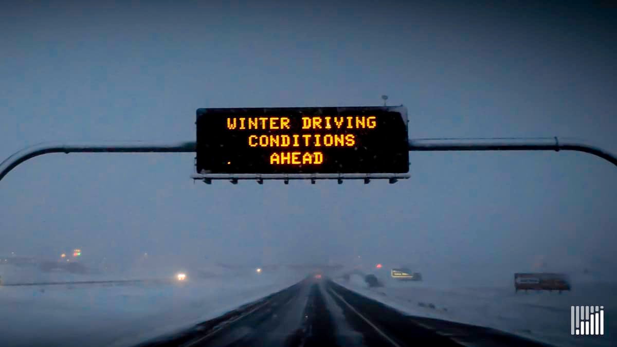 "Winter Driving Conditions Ahead" sign above a snow-covered highway.