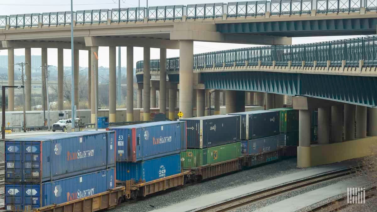 A photograph of a train with intermodal containers traveling underneath a bridge.