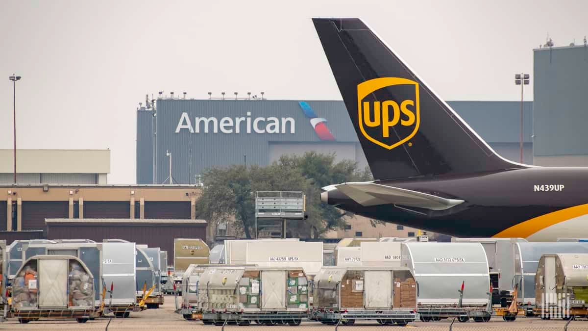 Brown tail of UPS jet in foreground, cargo containers and American Airlines cargo warehouse in background.