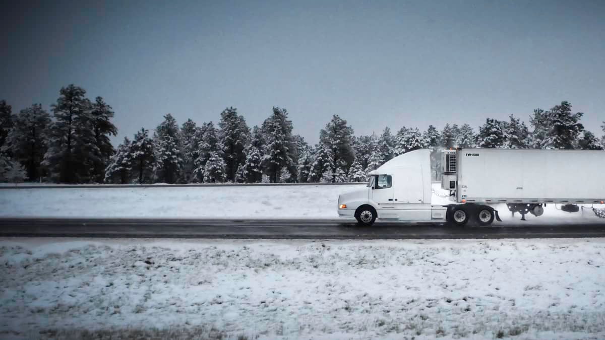 Tractor-trailer heading down highway with some snow on the road and surrounding hillsides.