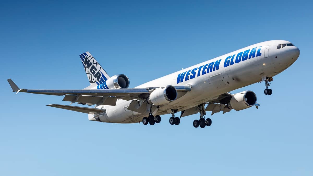 A white jet with three engines comes in for landing with wheels down against blue sky.