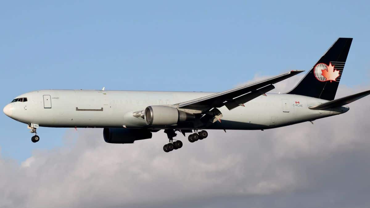 A white jet, with blue tail and Canadian logo comes in for landing with wheels down and a cloud in the background.