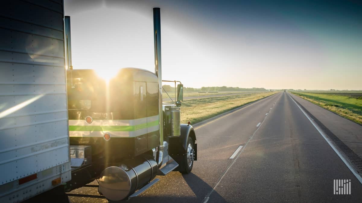 Tractor-trailer heading down a highway with bright sunshine on the horizon.