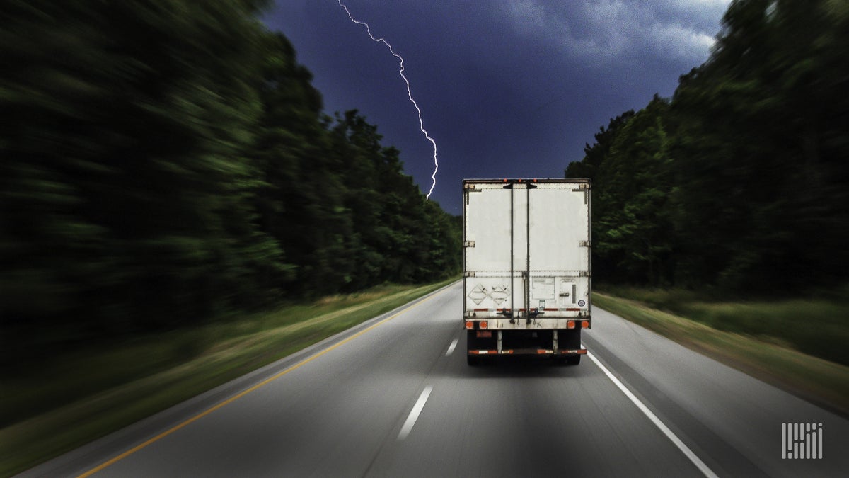 Tractor-trailer heading down a highway with lightning across the sky.