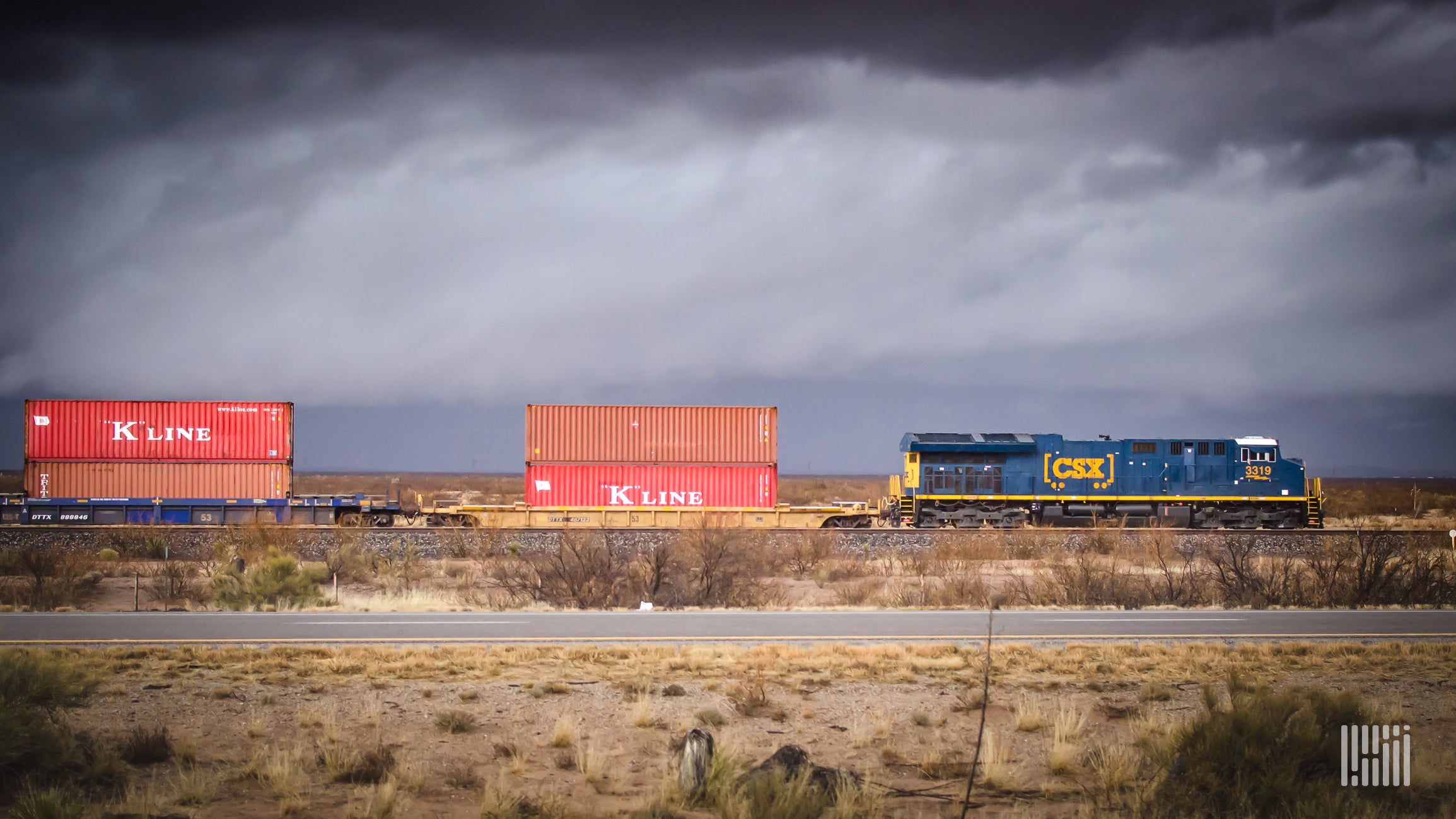 A photograph of a CSX intermodal train rolling through a desert field.