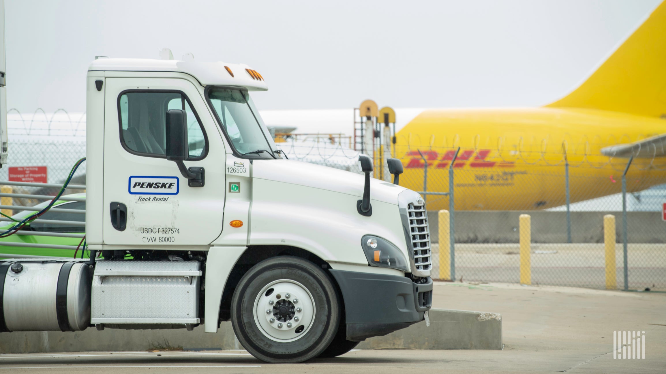 Back half of a mustard-colored DHL plane in background and tractor-trailer cab in foreground at airport.