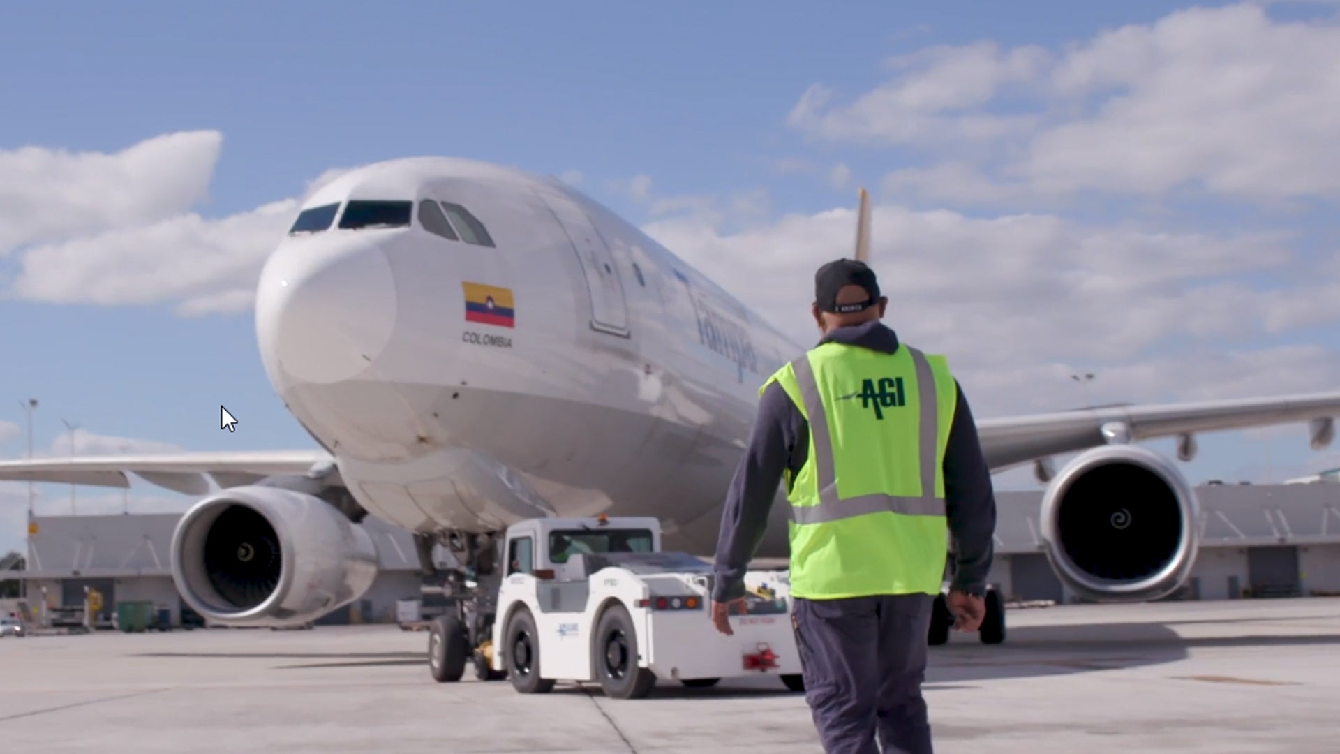 An airport worker with a yellow vest walks towards an airplane and a ground tug.