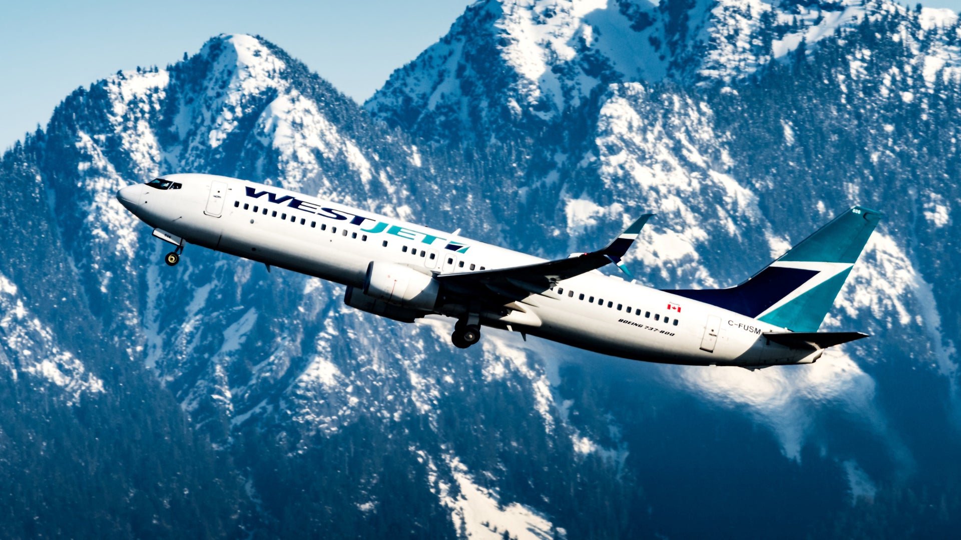 A white WestJet plane axcends with snow-covered mountains as backdrop.