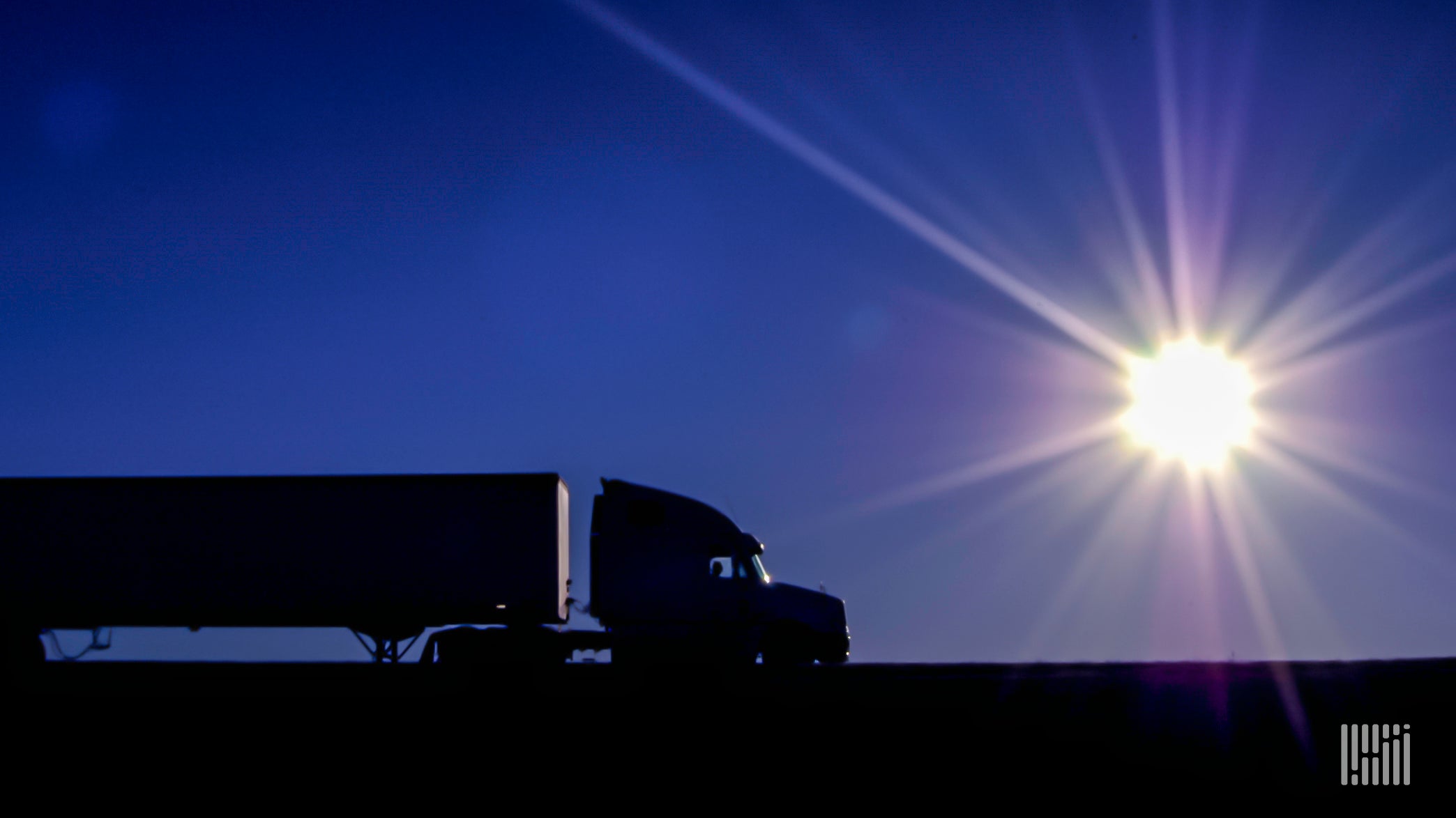 Tractor-trailer heading down a desert highway with scorching sun in the sky.