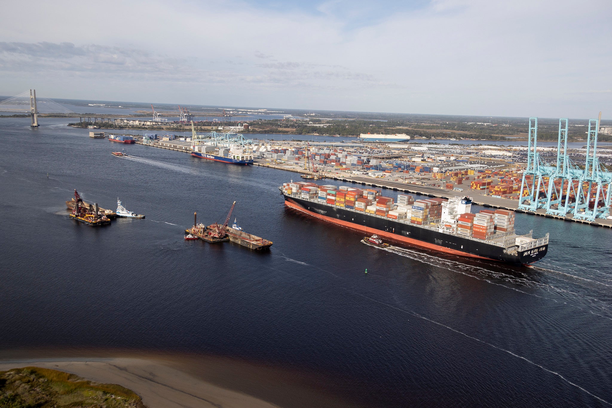 A photograph of a container vessel passing through a shipping channel. There are two smaller boats next to the vessel.