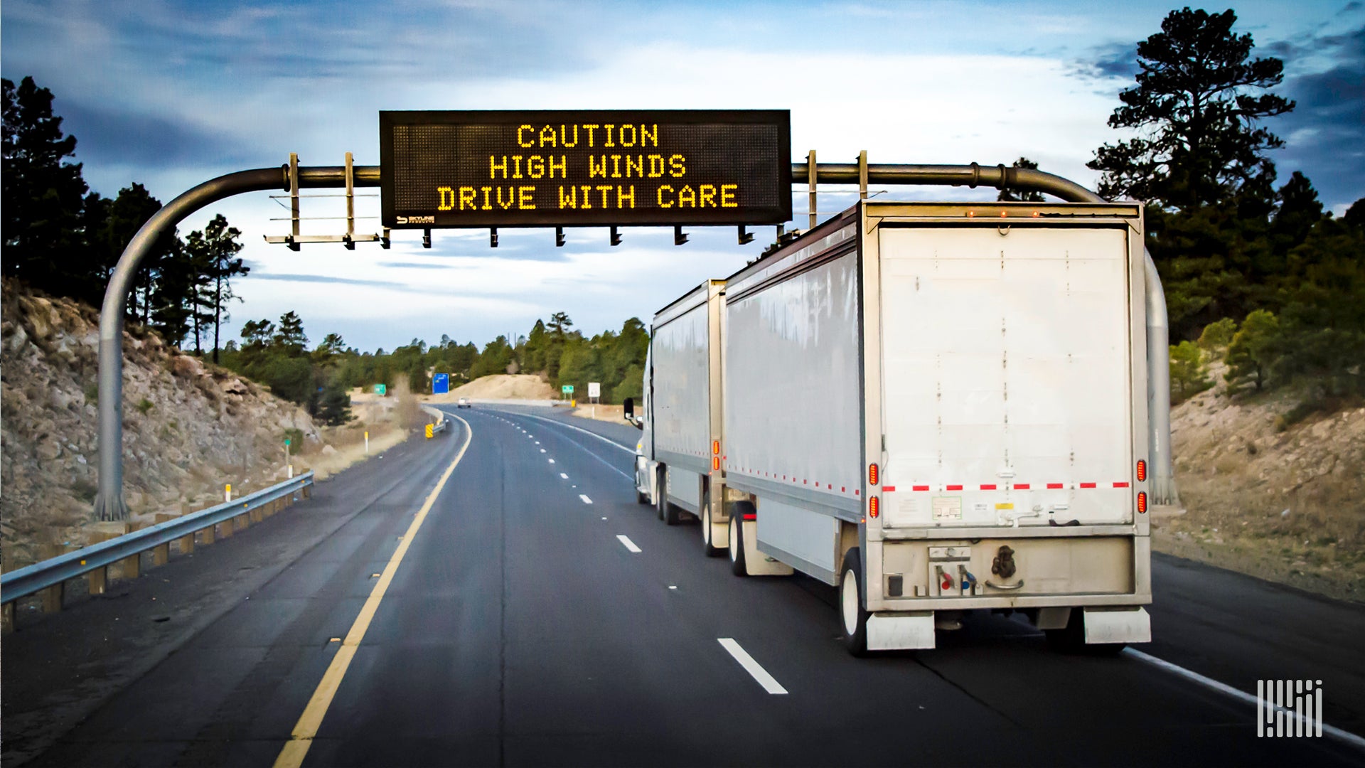 Tractor-trailer with "Caution High Winds" highway sign ahead.