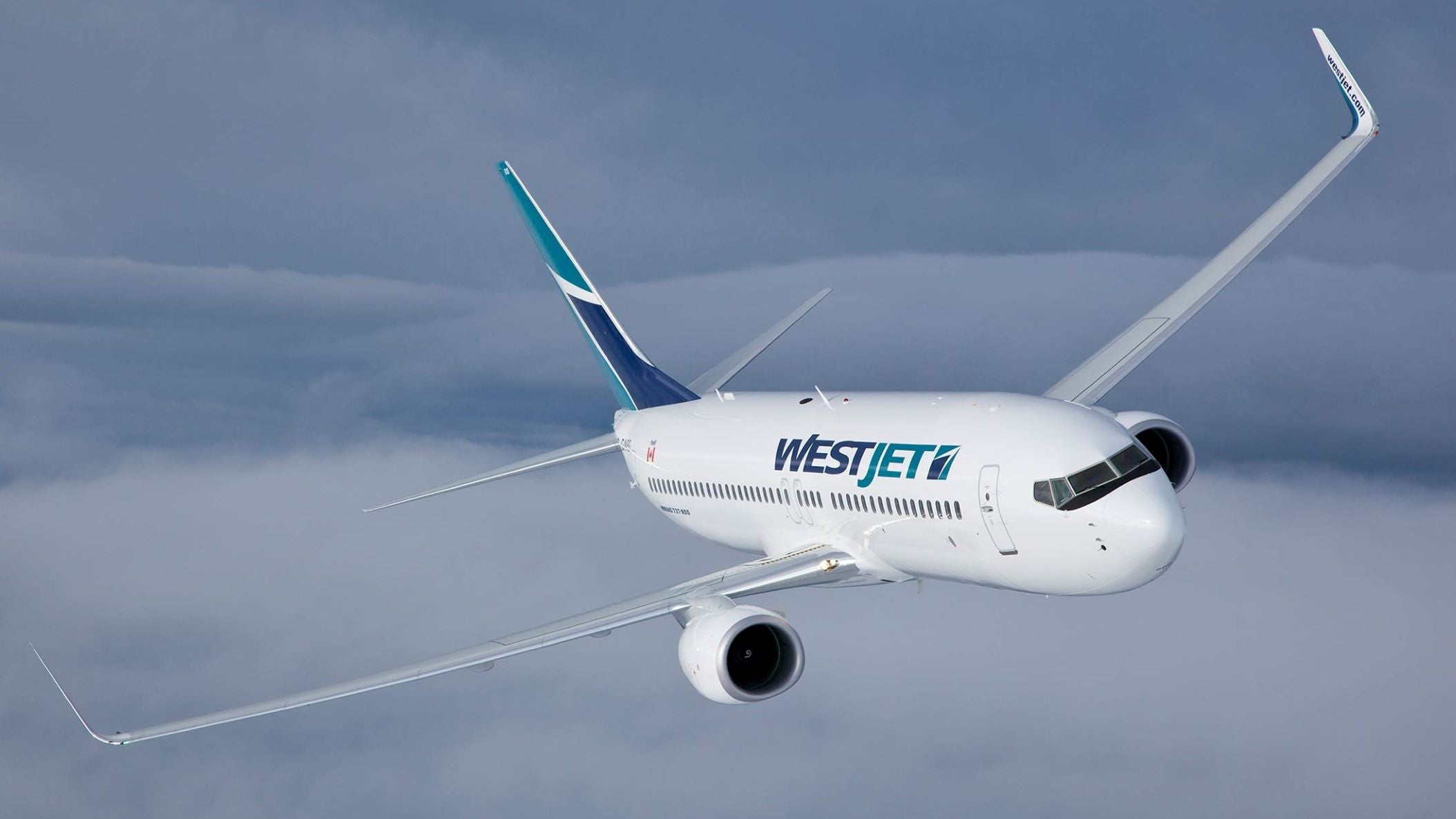 A white WestJet plane with aqua trim flying above the clouds and coming towards camera.