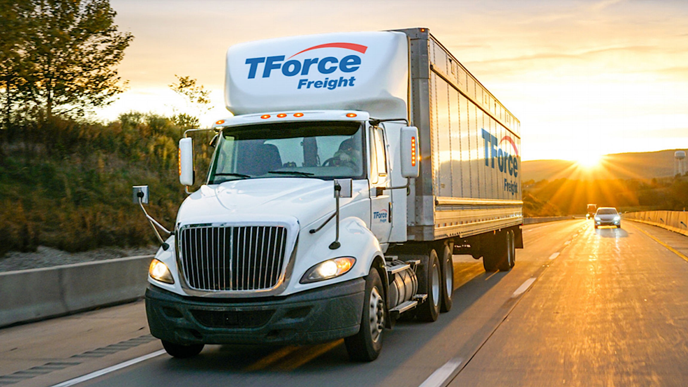 A white tractor-trailer TFI International carrier TForce Freight seen from the front travels on a highway as the sun sets behind it.