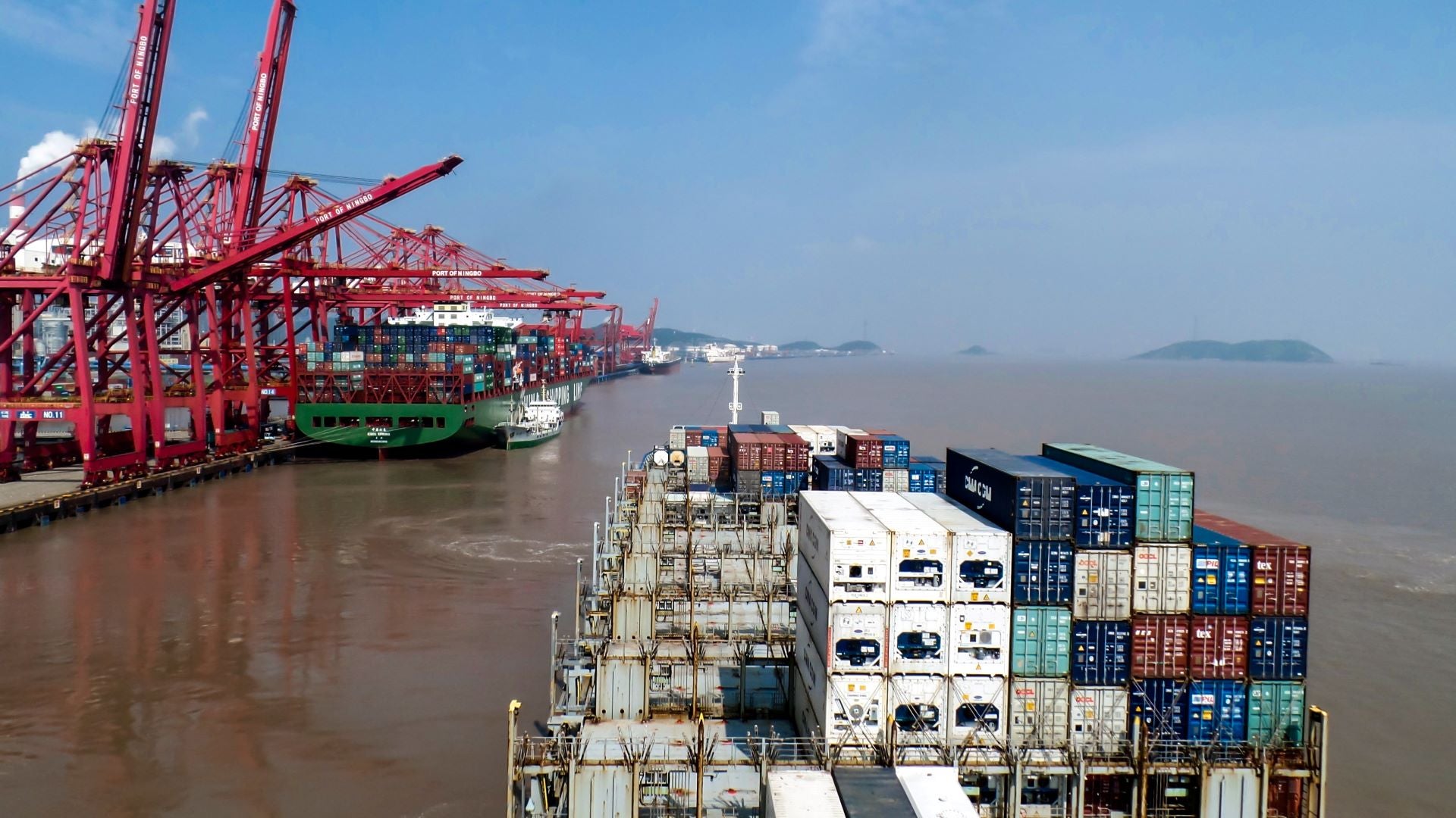 A view from the back of a container vessel entering a port with tall cranes on the dock.