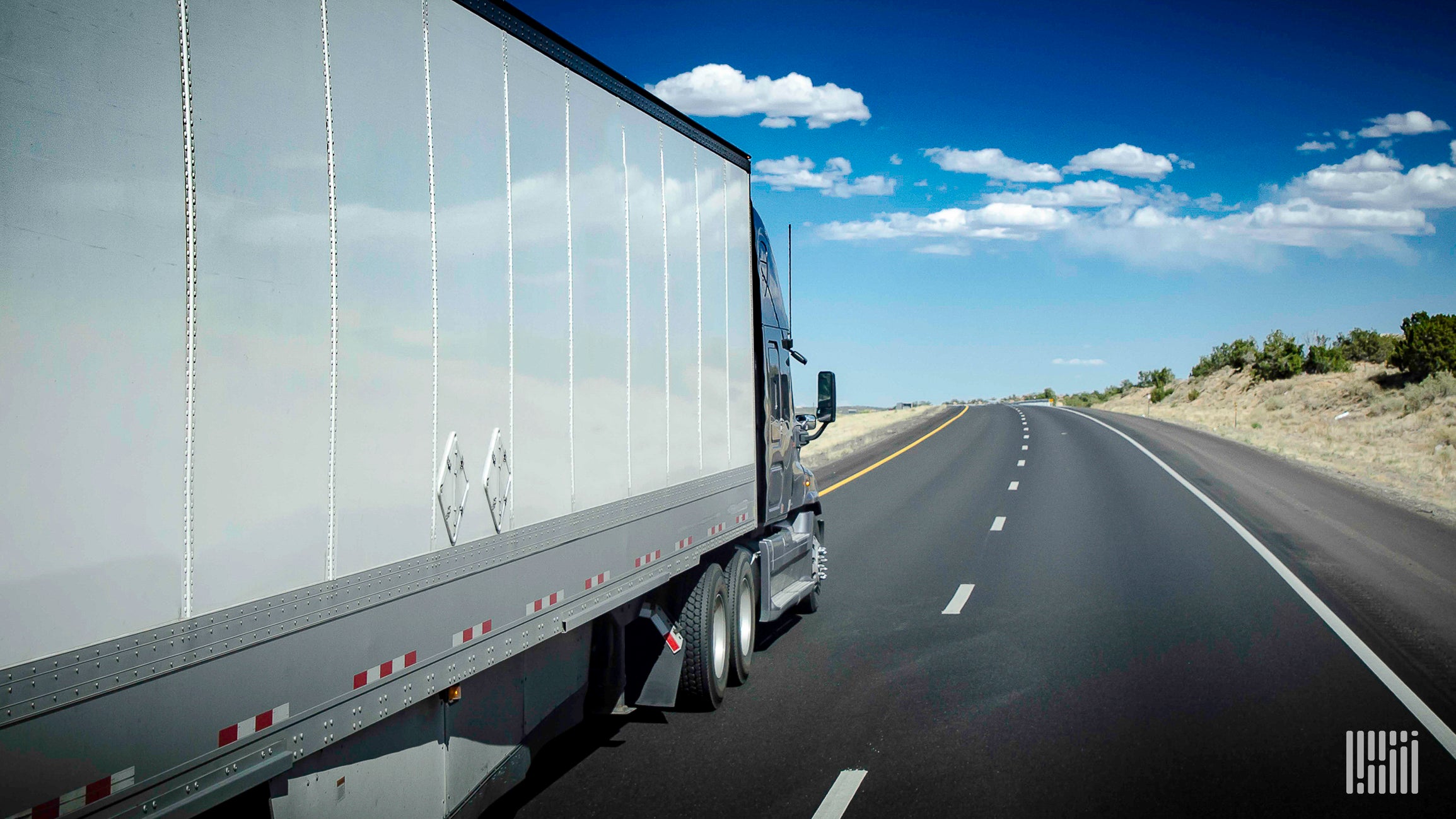 A white tractor-trailer travels on a highway viewed from the rear and two the right.