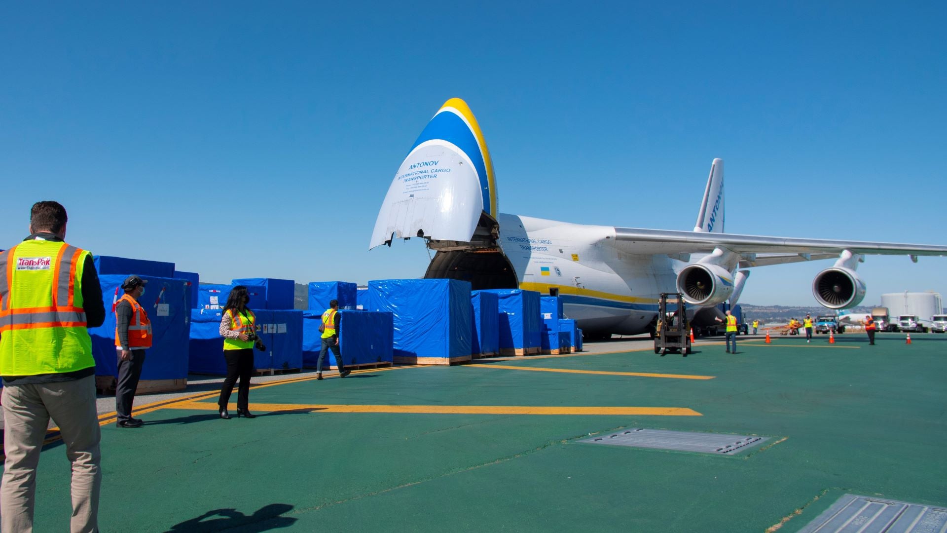 Large crates covered with blue tarps lined up in front of a nose-loading air cargo jet on a clear blue day.