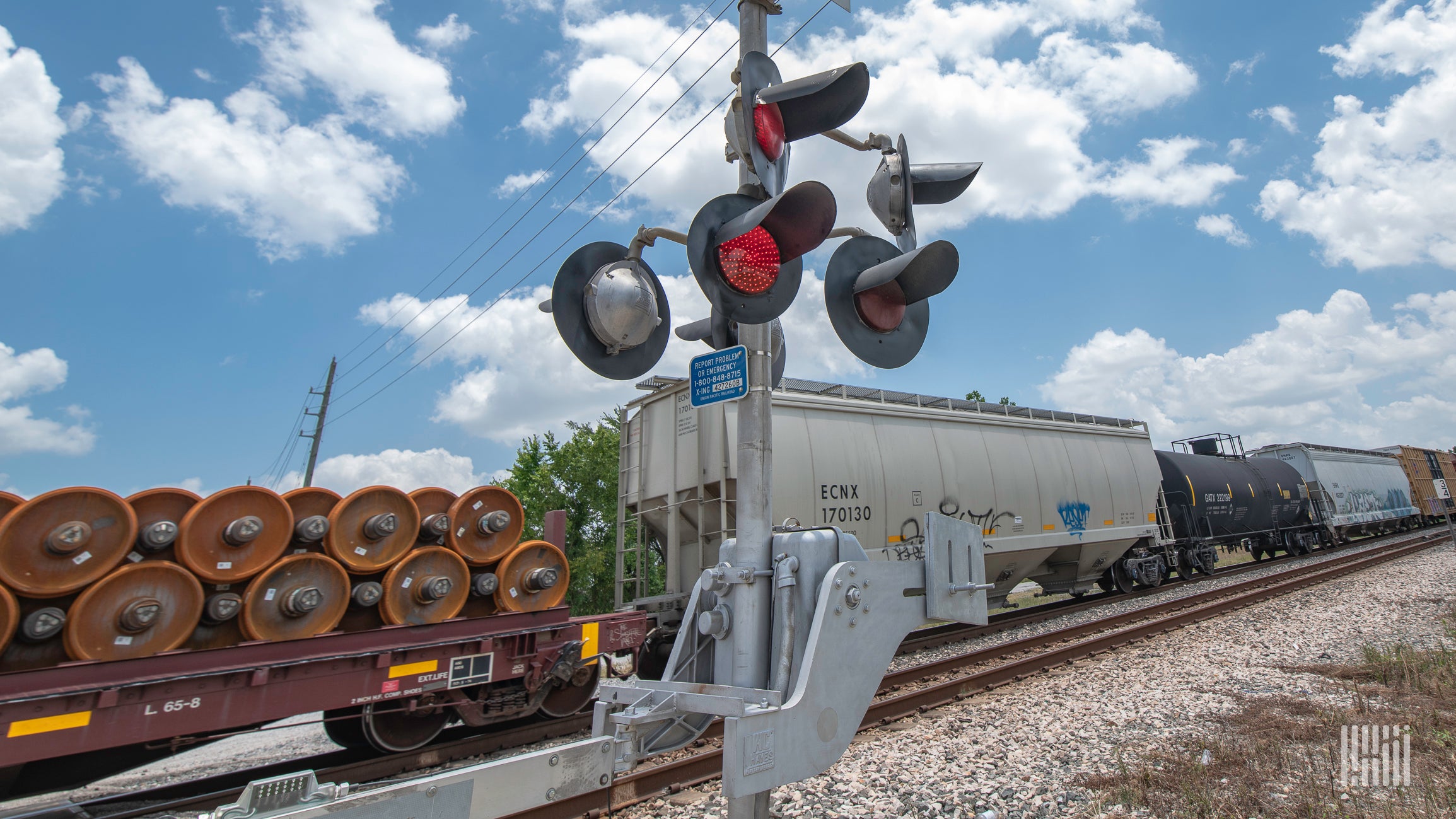 A photograph of a train passing by a rail crossing on a sunny day.