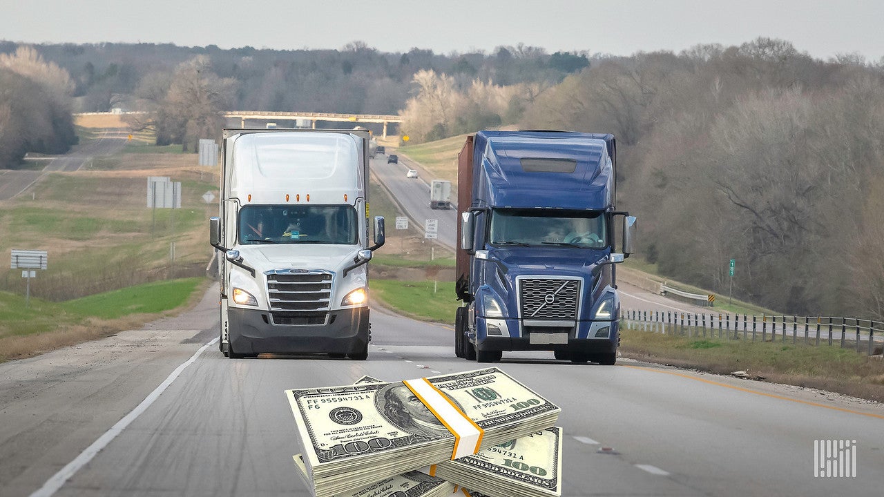 Two tractor-trailers, one white and one blue, travel next to each other on the road, where an image of a bundle of $100 bills is seen.