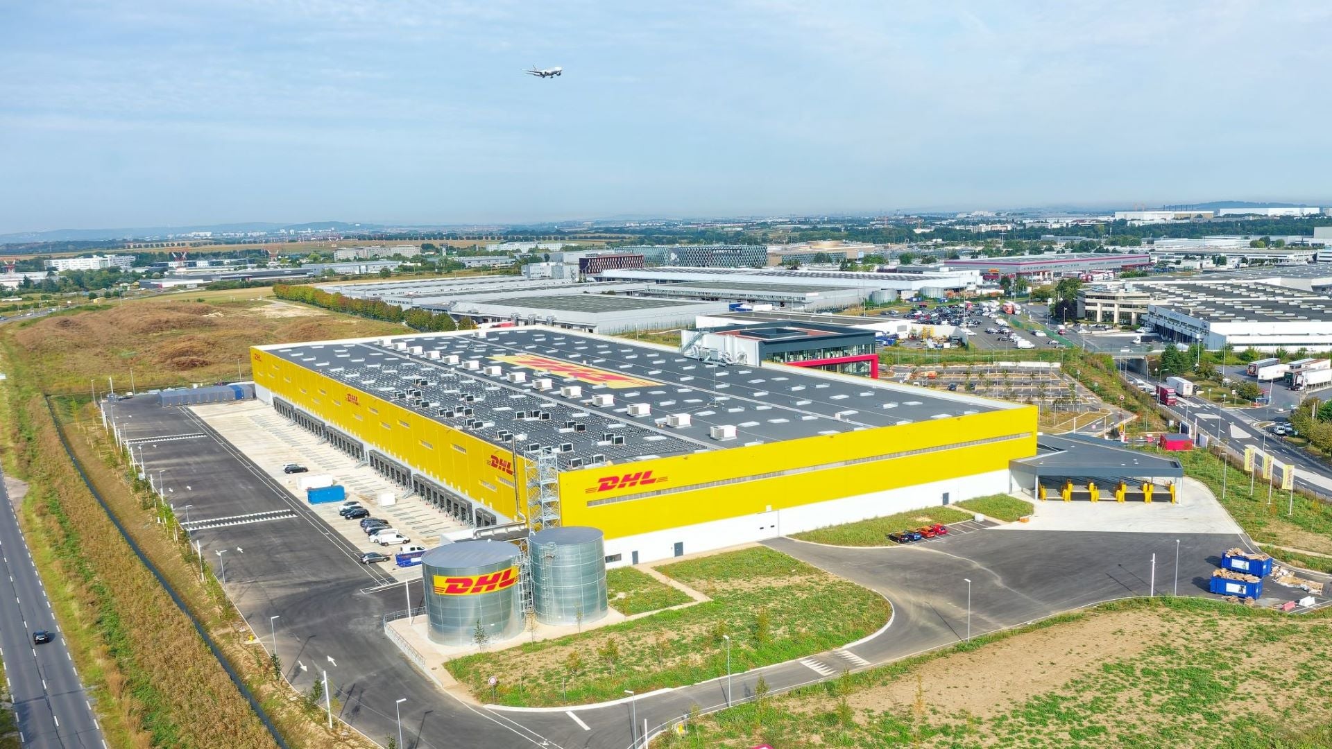 A big yellow warehouse with the DHL logo on the roof, looking down from above with an aircraft approaching in the distance.