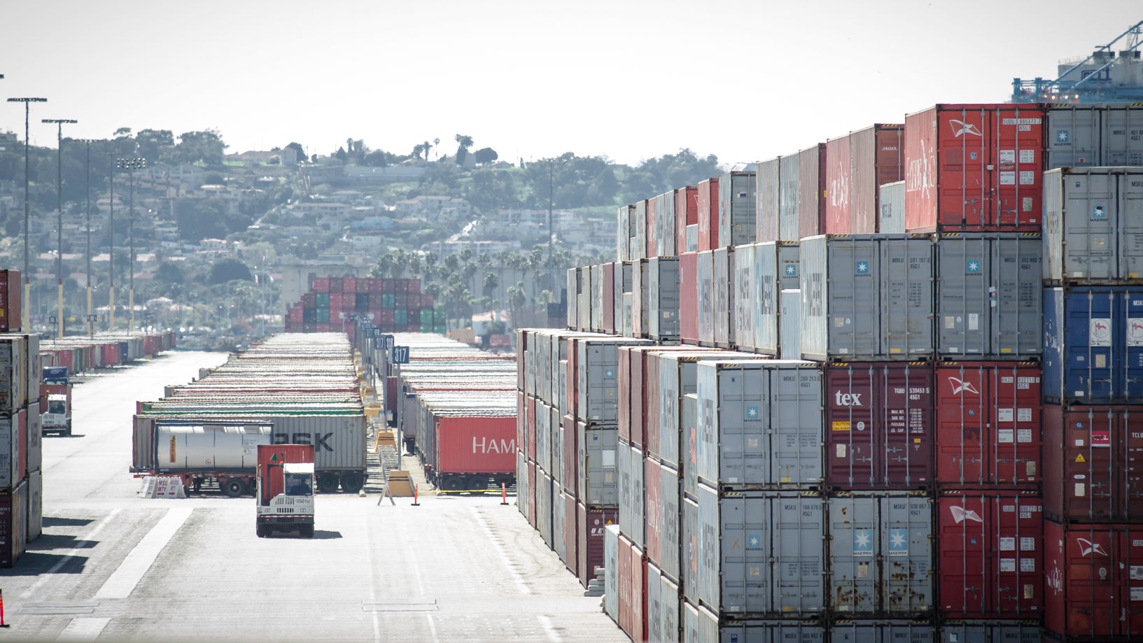 Stacks of containers at a port, with trucks in the background.